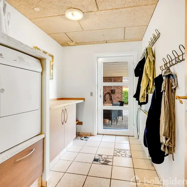 Vaskemaskine og tørretumbler er placeret på en bordplade. Tøj hænger på knager langs væggen. Rummet har flisegulv og en glasdør åbner ud til en terrasse.
