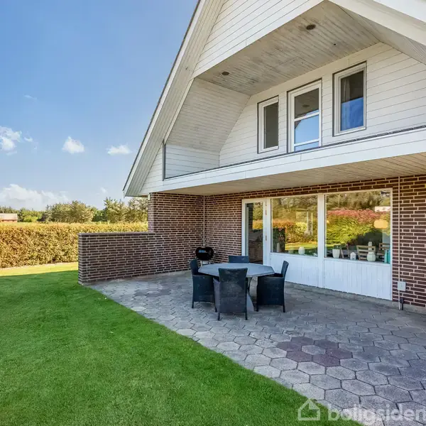 Et moderne hus med hvidt, skrånende tag og store vinduer står stille. Foran huset er der en flisebelagt terrasse med sorte havemøbler. Omgivet af en velholdt grøn græsplæne.
