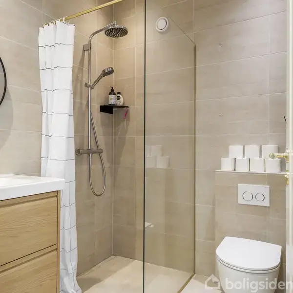 Badeværelse med bruseområde og glasvæg. Rundt spejl over vask med dispenser. Hvide fliser og moderne armaturer. Toilet til højre. Organisering med hylder. Lyst og funktionelt design.