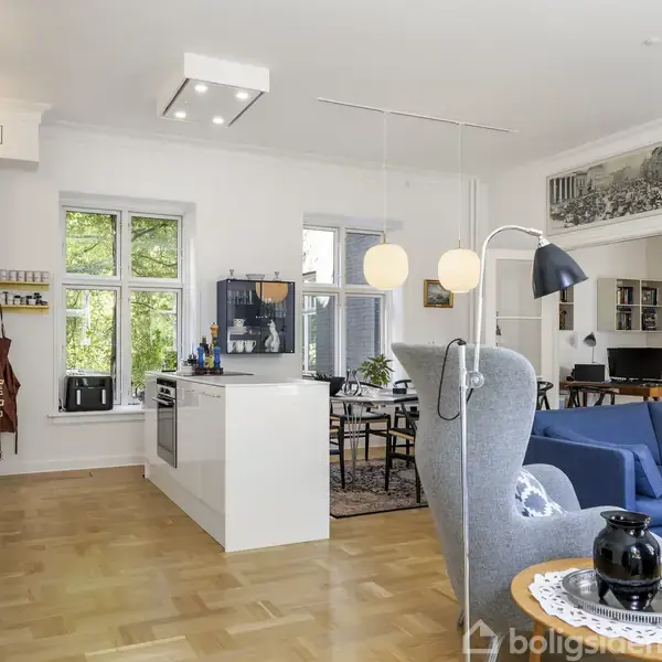 Lyst, åbent opholdsområde med moderne køkken, hvide skabe og køkkenø, stue med blå sofagruppe, designstol, og spisebord. Trægulv, store vinduer med udsigt til grønne træer, skaber lysfyldt atmosfære.