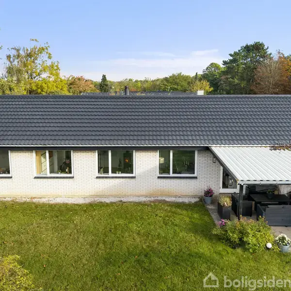 Huset står stille; en lav murstensbygning med sorte tagsten. Foran er der en velholdt græsplæne og en overdækket terrasse til højre. Omkranset af træer skaber en fredfyldt atmosfære.