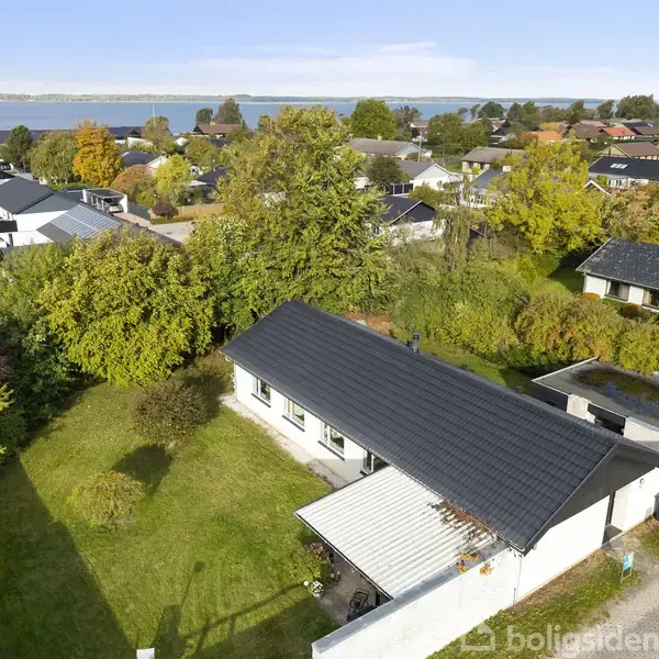 Huset ligger i en grøn have med tæt buskads. Omgivelserne har flere huse og træer, med en sø i horisonten. Solskinsvejr understreger den fredfyldte atmosfære.