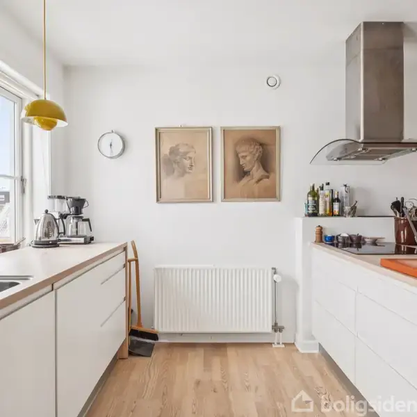 Et lyst køkken med hvide skabe og en vask til venstre, en emhætte og madlavningsredskaber til højre. Væggene har portrætter og en ur.