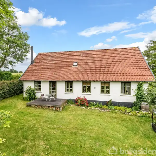 Et hvidt hus med rødt tag står stille i en frodig have med græsplæne og træer omkring. En lille terrasse med havemøbler ses foran huset.