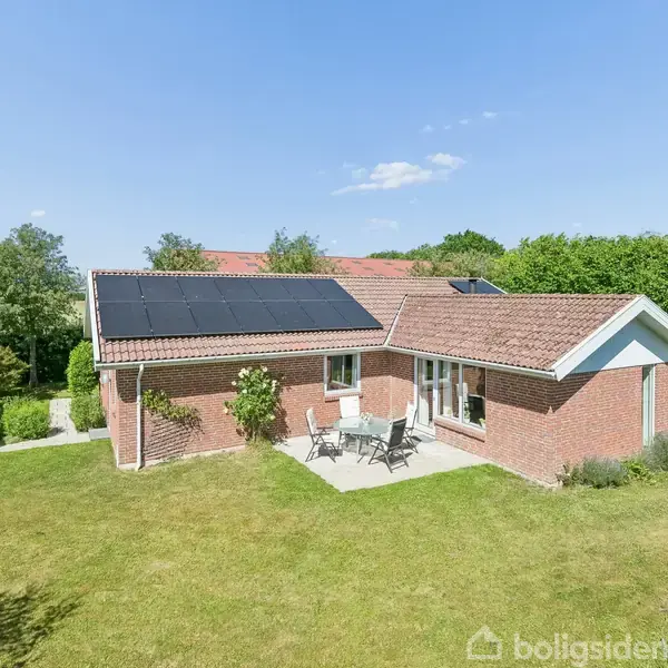 En rød murstensvilla med solpaneler på taget står i en grøn have med græsplæne. Foran huset er der en terrasse med havemøbler. Skyfri himmel i baggrunden.