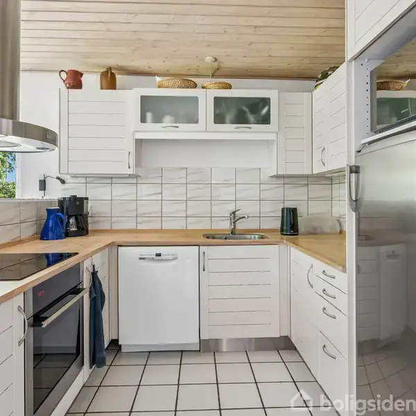 Køkken med hvide skabe og stål apparater. Kaffemaskine på bordplade. Fliser på vægge, trægulv. Lysindfald fra vindue, planter udenfor. Hyggelig og moderne indretning. Skinnende rent.