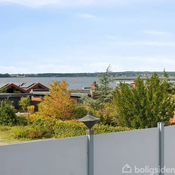 Et hegn står foran grønne træer og buske i en forstadshave med udsigt over en sø samt huse i baggrunden under en blå himmel.