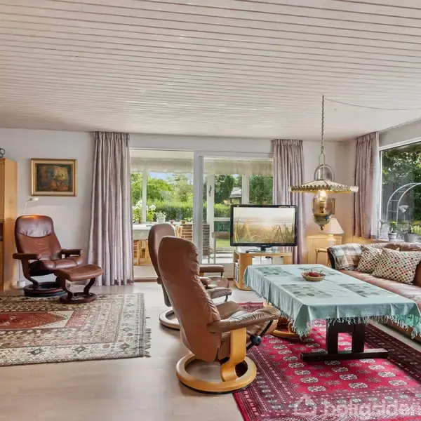 Stue med lædersofa og stole omkring et sofabord med blå dug. Store vinduer viser en have. I baggrunden står en trækommode, og et tæppe dækker gulvet.