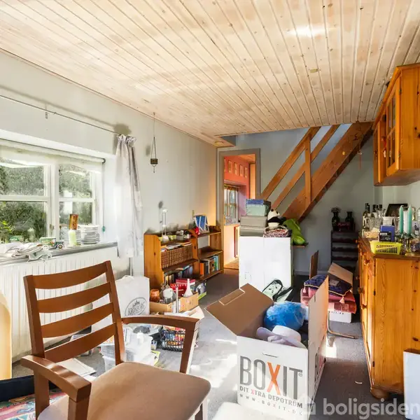 Et rum er fyldt med forskellige møbler og kasser. Aktion: Kasser med mærket "BOXIT" står midt i rummet. Kontekst: Træloft, en trappe og et stort vindue i en lys stue.