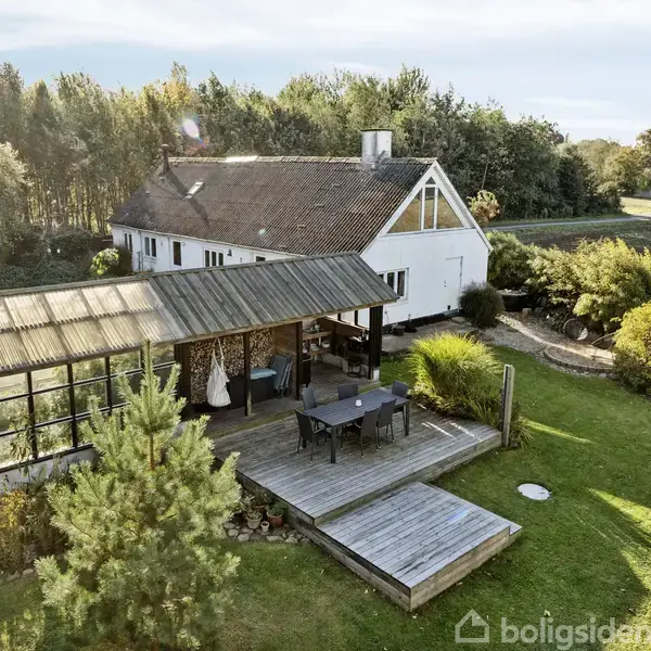 Hus i hvidt med mørkt tag. En overdækket terrasse med spisebord ses foran, omgivet af grøn have og træer. Landlig beliggenhed med marker i baggrunden.