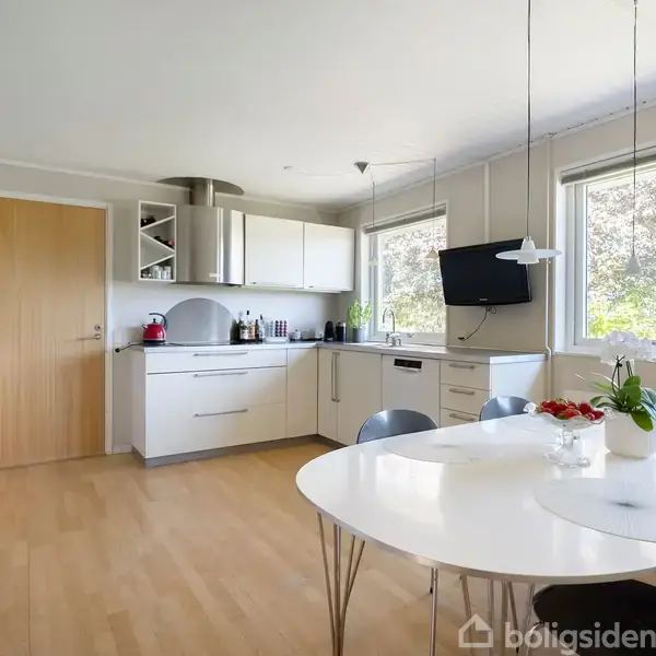 Køkken med hvidt bord og stole, omgivet af lyse træskabe og apparater. Vinduer lukker lys ind, mens en fladskærms-tv hænger på væggen. Bordet har planter og frugter.