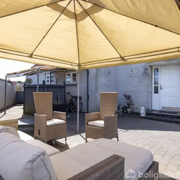 En terrasse har møbler under en beige pavillon. Møbleringen består af en sofa og to stole. I baggrunden ses en cykel op ad en grå, pudset mur ved en hvid dør.
