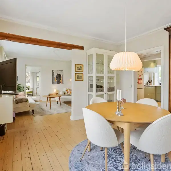 Et rundt spisebord med fire stole står i en lys, elegant stue. En stor spejl står til højre. Stuen har trægulv, tæpper og en TV i baggrunden. Planter dekorerer rummet.
