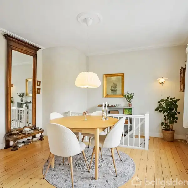 Et rundt spisebord med fire hvide stole står på et rundt tæppe i en lys spisestue med trægulv. Store vinduer og planter i potter skaber en hyggelig atmosfære.