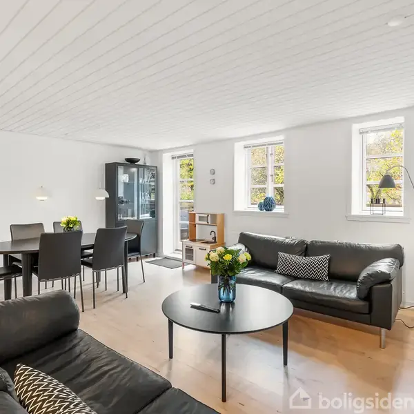 En moderne stue med en sort lædersofa og sofabord med blomster. Et spisebord med stole står ved siden af et vitrineskab. To vinduer giver lysindfald.