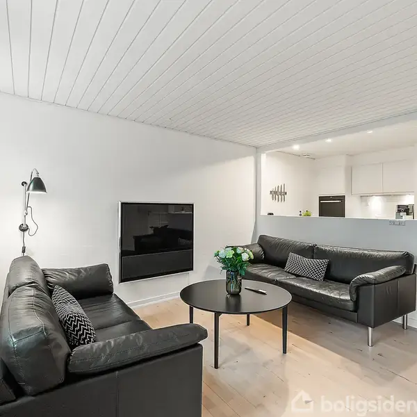 To sorte lædersofaer står i en stue med hvide vægge og lofter. En fladskærms-tv hænger på væggen. Rundt sofabord med blomster. Køkken ses i baggrunden.