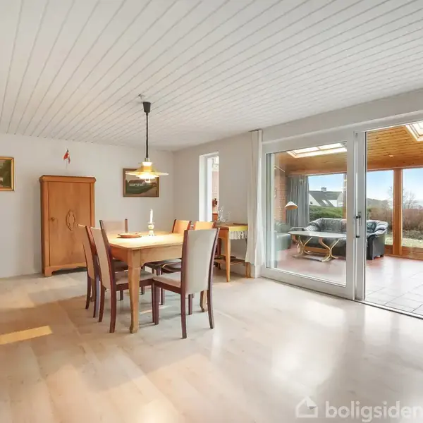 Rødstenshus med hvid træbeklædt carport foran. Nummeret "59A" vises tydeligt på træværket. Bygningen har små vinduer og er omkranset af blå himmel med få skyer.