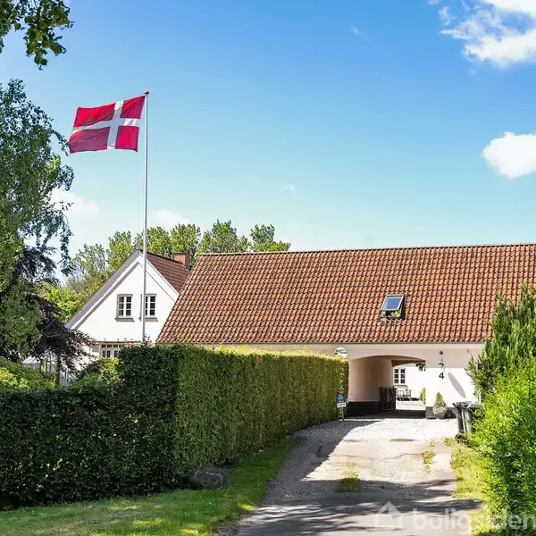 Et hvidt hus med rødt tegltag står omgivet af frodig vegetation. En dansk flagstang med Dannebrog vajer til venstre. Blå himmel med spredte skyer danner baggrund.