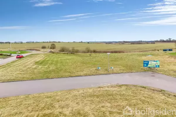 En rød bil kører på en landevej omgivet af åbne, grønne marker under en klar, blå himmel. Et skilt på højre side angiver vej og mulig ejendom.