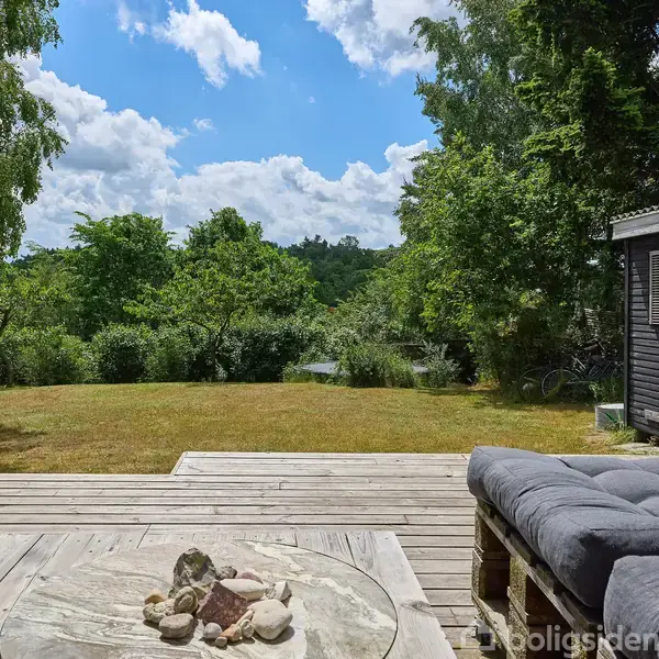 Sofa står på en træterrasse med udsigt over en græsplæne og frodig have. Et lille træskur står til højre. Himlen er blå med hvide skyer i baggrunden.