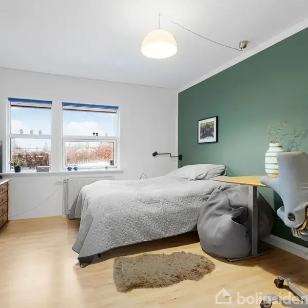 Soveværelse med seng dækket af gråt sengetæppe, stående lamper over natbord. Til højre et skrivebord med kontorstol og computer. Grøn væg bagved, vinduer med gardiner og landskabsudsigt.