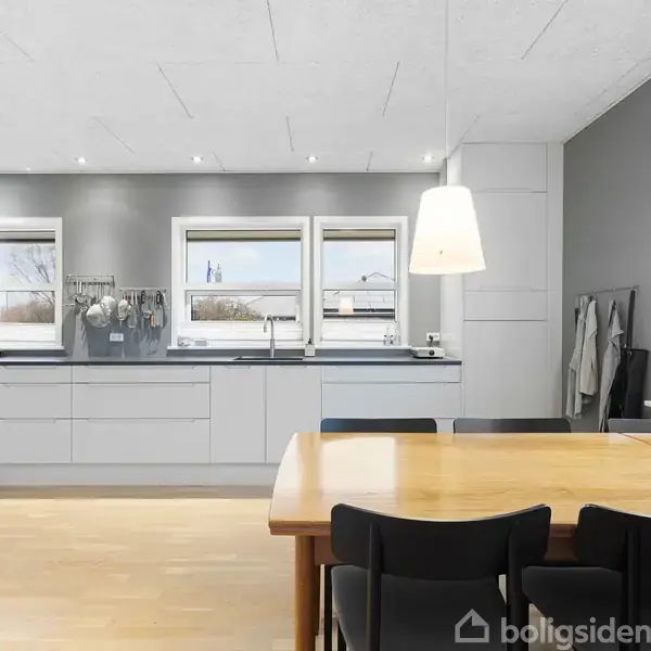 Et moderne køkken med hvide skabe er indrettet langs væggen. Et træbord omgivet af sorte stole står i forgrunden. Store vinduer giver udsigt til udendørs omgivelser.