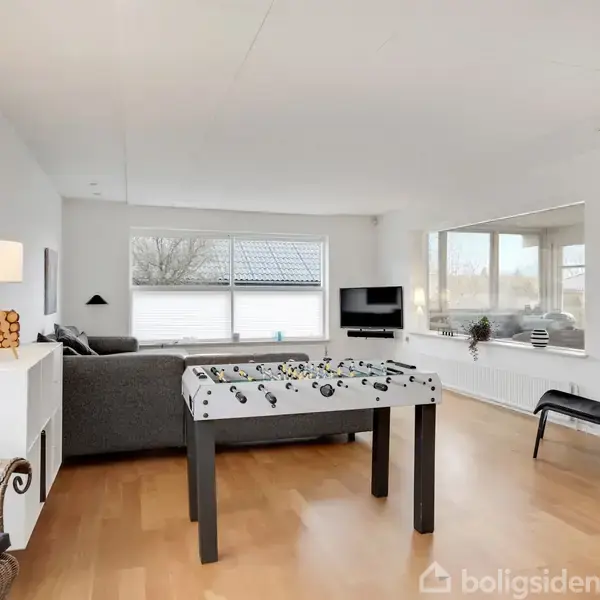 Et bordfodboldbord står i en stue med parketgulv. Stuen har en grå sofa, fladskærms-tv på væggen, og store vinduer med udsigt til en gårdhave.