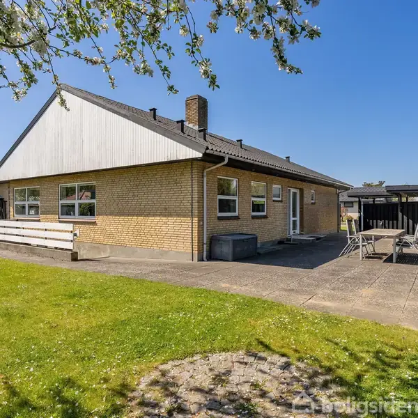 Hus lavet af gule mursten står stille på en solbelyst terrasse omgivet af græsplæne og træer. En sort trævæg og havemøbler ses til højre.