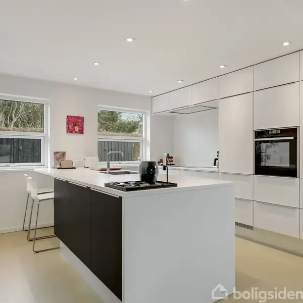 Et moderne køkken med en central køkkenø udstyret med en kogeplade, omgivet af hvide skabe og indbygget ovn. Lyse vægge, store vinduer og minimalistisk indretning skaber et rummeligt miljø.