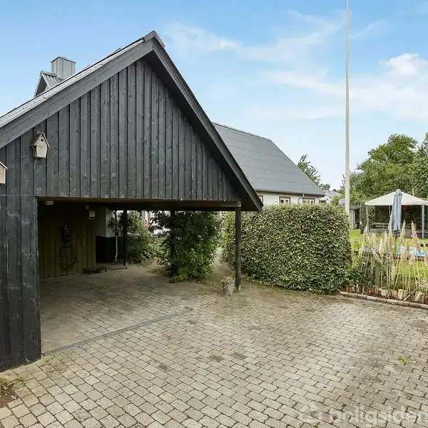 En sortmalet carport står stille på en brolagt indkørsel omgivet af hække. En havepavillon med parasol ses i baggrunden, og omgivelserne har frodige træer og buske.