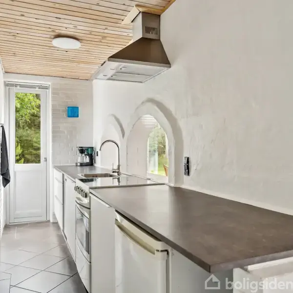 Et smalt, moderne køkken med en lang bordplade, ovn og emhætte; lysindfald fra dør og vinduer. Vægge er hvidmalede med synlige mursten og loftet er træbeklædt.