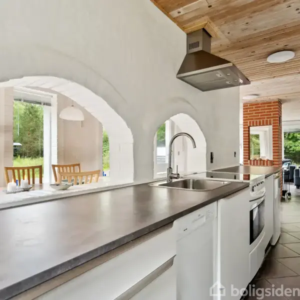 Køkkenbord med indbygget ovn og emhætte, flankeret af buede åbninger. Bagved ses en spisestue med vinduer mod en grøn have samt en stue med sofa.