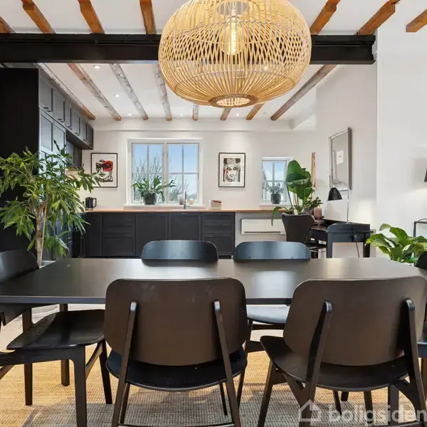 Spisebord med seks sorte stole er placeret under en stor, flettet lampe. Køkkenet har sorte skabe og masser af grønne planter. Væggene er hvide, og loftet har synlige bjælker.