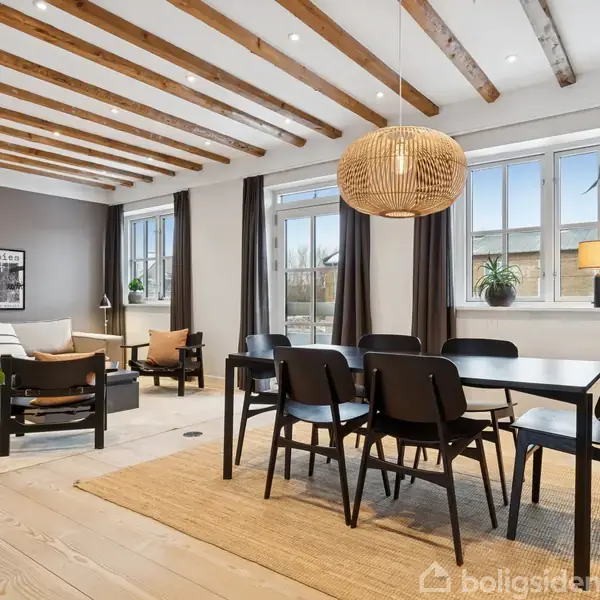 Spisestue med sort spisebord og seks stole, overskygget af en strålampe, placeret på et tæppe. Loftet har synlige træbjælker. Værelset har store vinduer og indendørs planter.