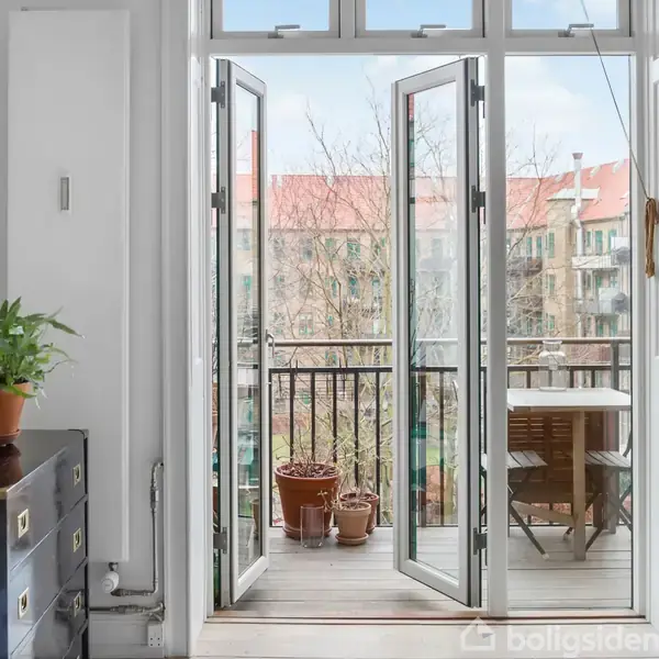 Franske døre åbner ud til en altan med planter og et bord-stolesæt. Inde i rummet ses en sort kommode med en lampe og en plante.