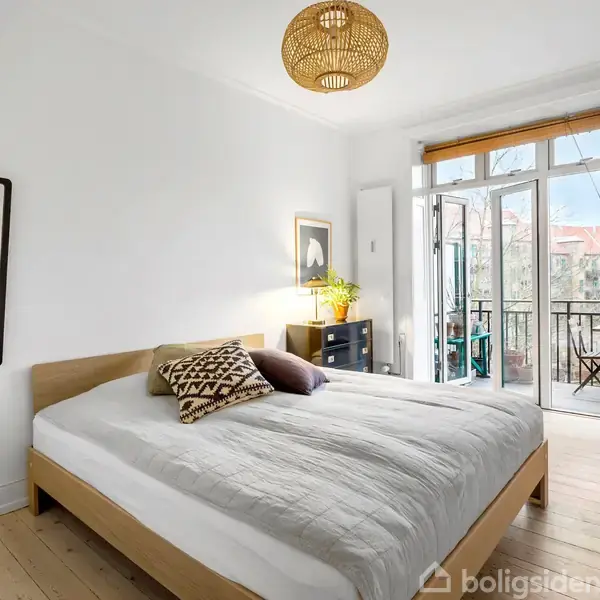 En dobbeltseng med puder står i et lyst soveværelse. Rummet har trægulv, en kommode med en plante, en vægmonteret spejl, og en udgang til en balkon med glasdøre.