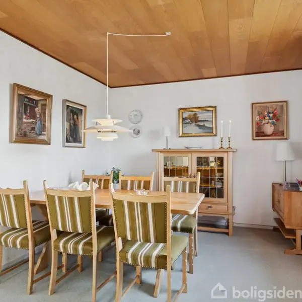 Et spisebord med stribede stole står under en pendellampe. Rummet har træloft, ophængte malerier og et vitrineskab med lysestager. Ved siden er en reol og vindue med røde gardiner.