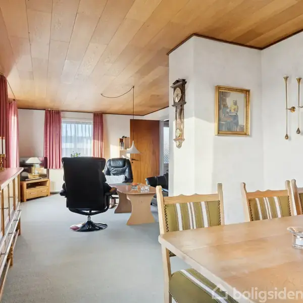 En stue med spisebord og stole står til højre. I baggrunden er der lænestole og et sofabord ved vinduer med røde gardiner. Rummets vægge er dekoreret med billeder.