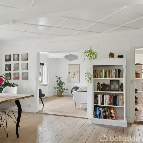 En lys stue med trægulv indeholder et spisebord med blomster samt en reol med bøger. Rummet åbner til en stue med sofaer og til et køkken med køkkenskabe.