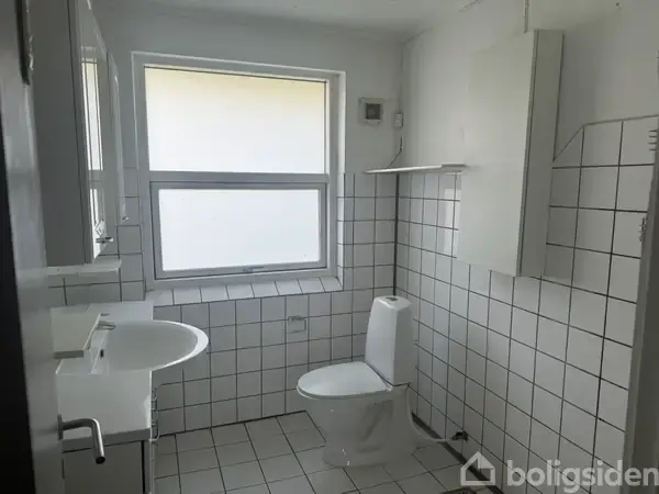 Toilet står stille i et hvidt, fliset badeværelse med en håndvask i nærheden og et vindue, der giver naturligt lys.