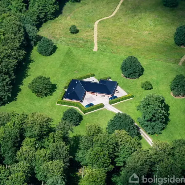 Hus med sort tag står på en stor græsplæne omgivet af træer og stier.