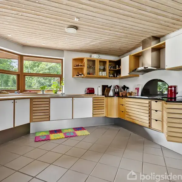 Køkken med træskabe og hvide facader står mod en væg med store vinduer, der afslører en frodig udendørsudsigt. Gulvet har store lyse fliser, og der er farverige tæpper.