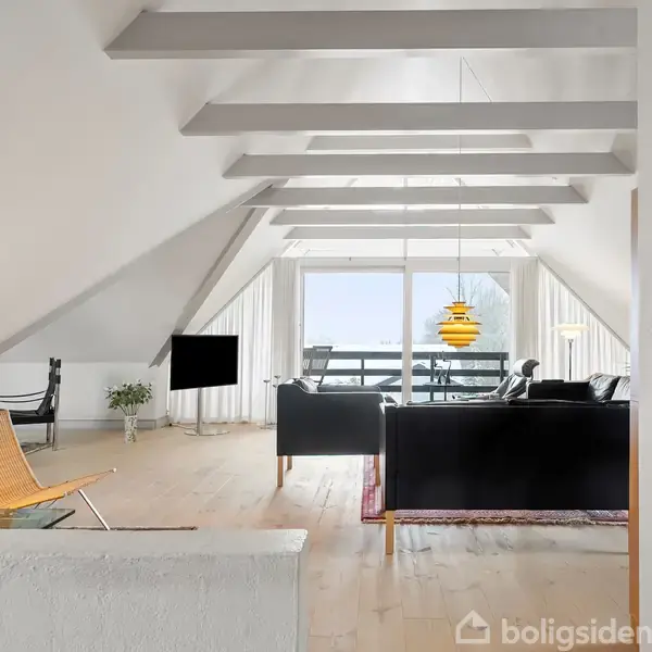 Stue med skrå lofter har en sort sofa og en gul lampe over et bord, placeret i en lys loftslejlighed med store vinduer og træmøbler. Telefon hænger på væggen.