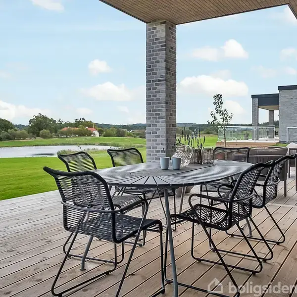 Terrassemøbler står ubrugte på en bred træterrasse med udsigt over en sø i en landlig baggrund. Terrasse er delvist overdækket af et moderne murstenshus med store vinduer i nærheden.