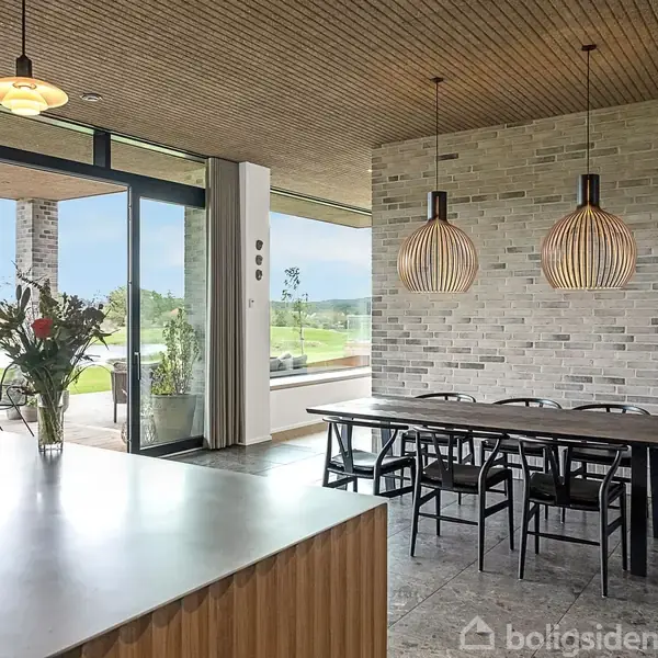 Moderne spisebord med sorte stole står under to hængelamper. Et stort vinduesparti åbner mod en terrasse og have. Indretningen har lyse murstensvægge og træloft. En køkkenø står i forgrunden.