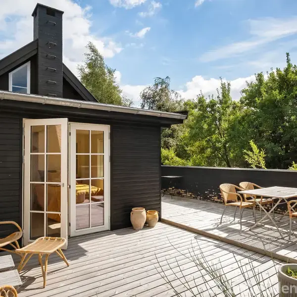 Et træterrasse med flettede stole og et bord i et frodigt, grønt skovområde. Franske døre åbner ind til et mørkt træhus under en blå himmel.