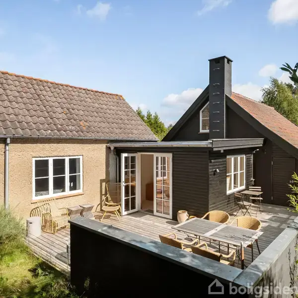 Hus i to dele med røde tegltage og sorte trævægge. Terrasse med borde og stole. Omgivet af grønne buske og træer under en blå himmel med få skyer.