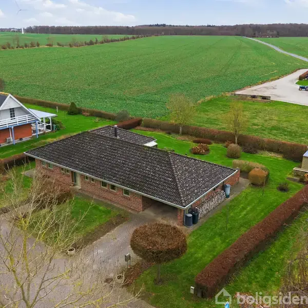 Et murstenshus med mørkt tag står i en have med velplejede buske. I baggrunden ses store grønne marker og en nabobygning. Der er ingen tekst i billedet.