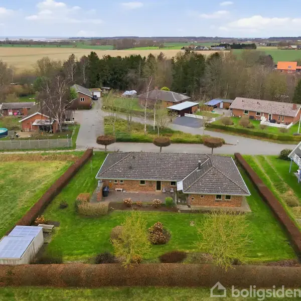 En murstensvilla beliggende i en velholdt have omgivet af hække. Omkringliggende huse og landevej ses i et naturskønt, landligt område med åbne marker og spredt skov i horisonten.