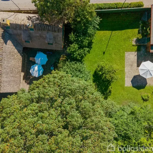 Stråtækt bindingsværkshus med gul facade, brun træstruktur og sort fundament. Huset står i en gårdhave med grønne buske, fliserenser og udendørs spiseområde under en blå, delvist skyet himmel.